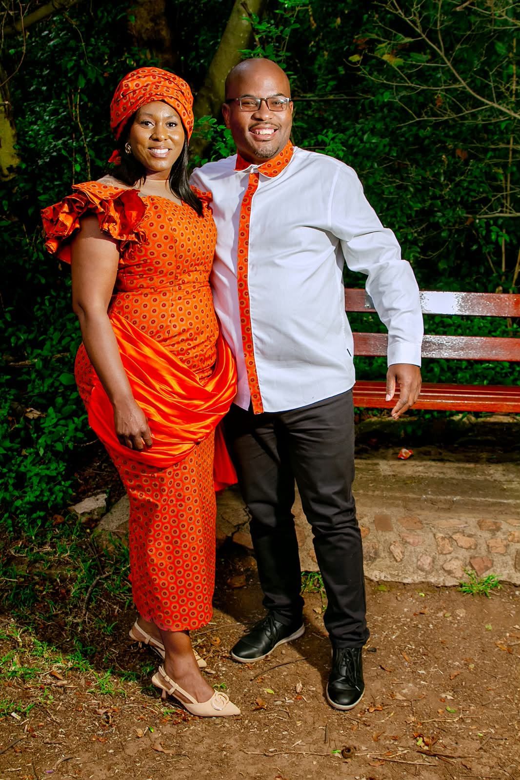 The couple standing in a garden in traditional attire, she in a fitted orange print dress with a satin drape and headwrap, he in a white shirt with orange print trim