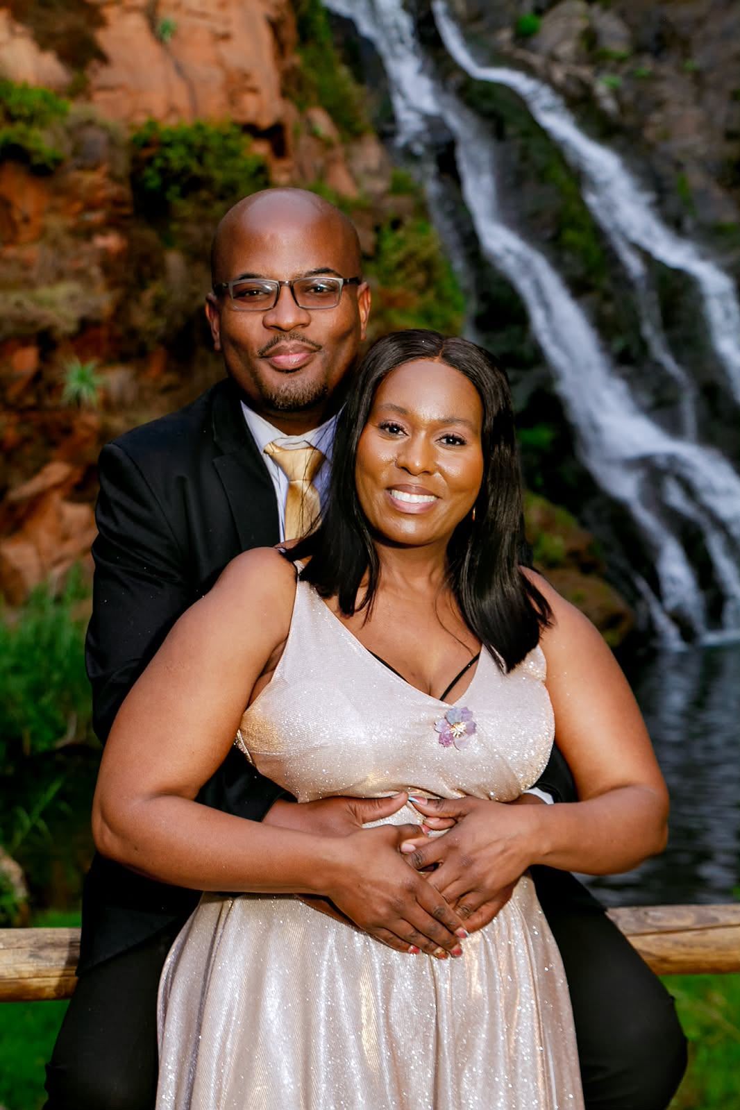 The couple in formal wear smiling, his arms wrapped around her waist, with a waterfall behind them