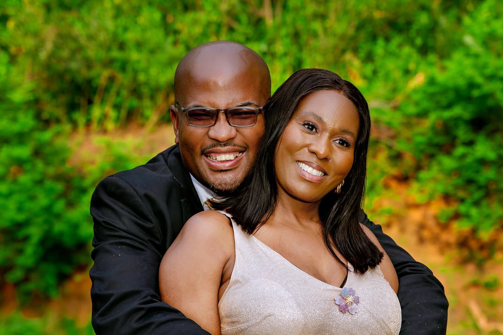 The couple laughing together as he hugs her from behind, he in a dark suit and she in a shimmering champagne gown