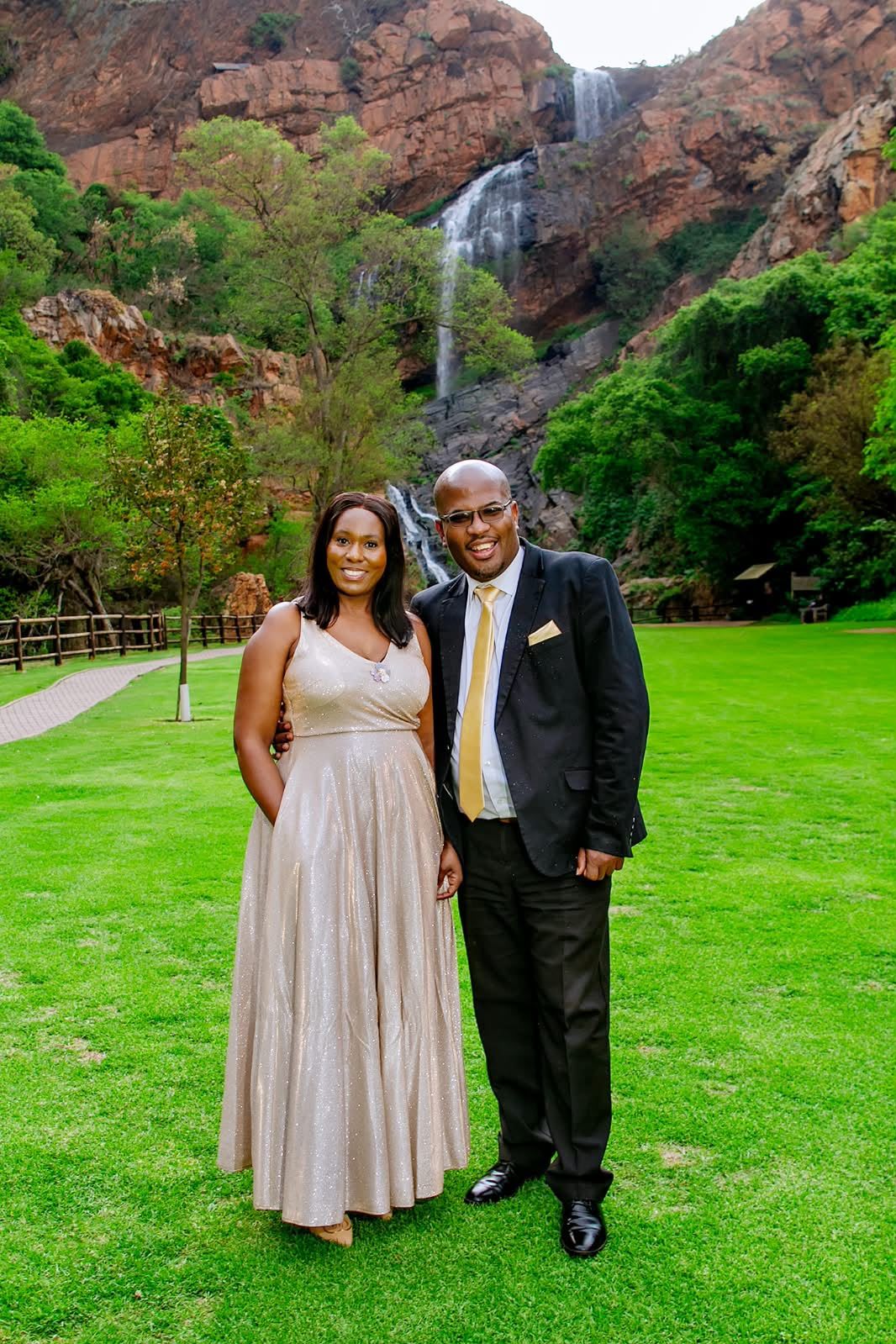 The couple in formal wear on a lawn below a waterfall, she in a floor-length champagne gown, he in a dark suit with a gold tie