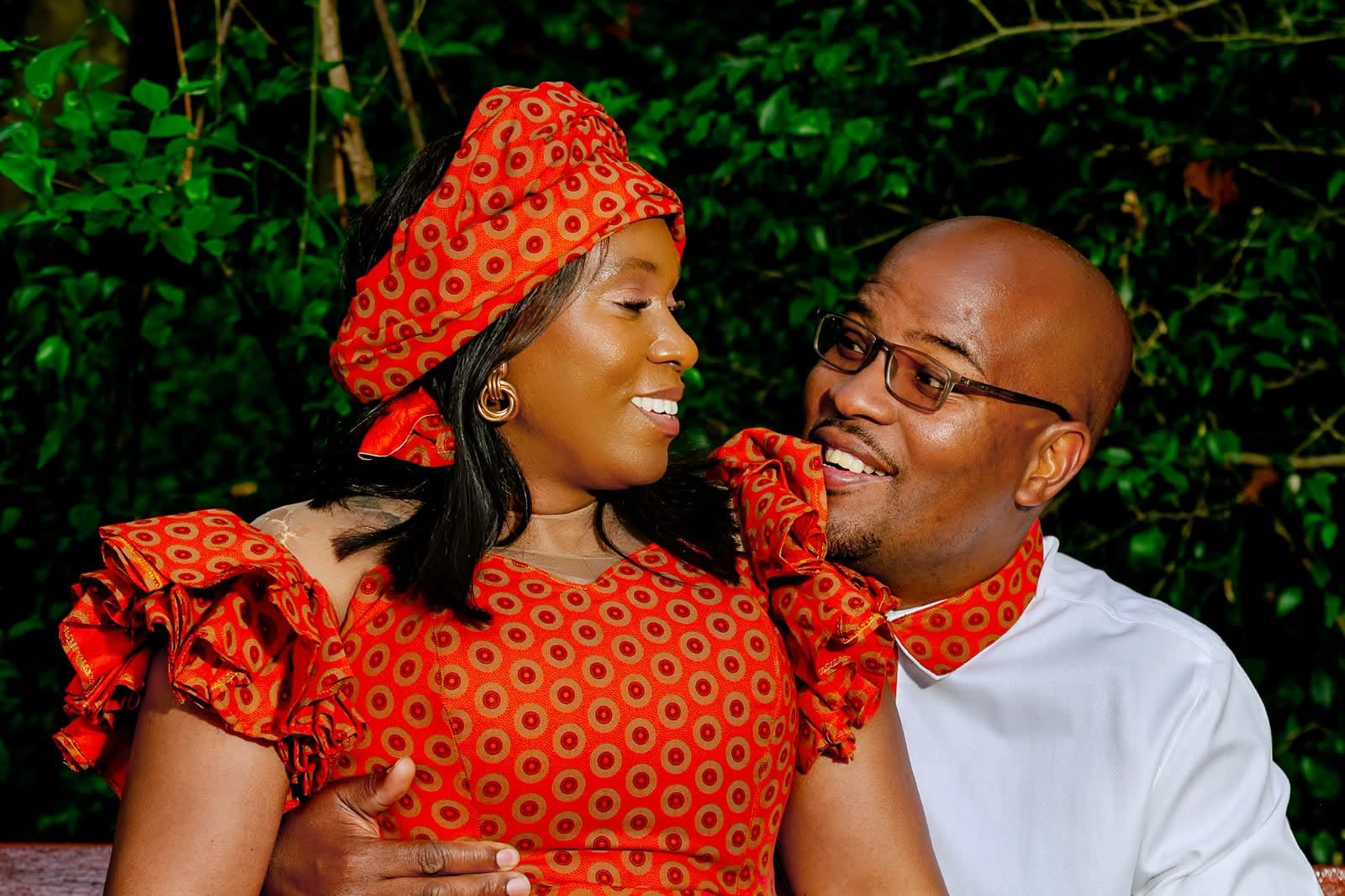The couple in orange traditional print attire smiling at each other in front of dark green leaves