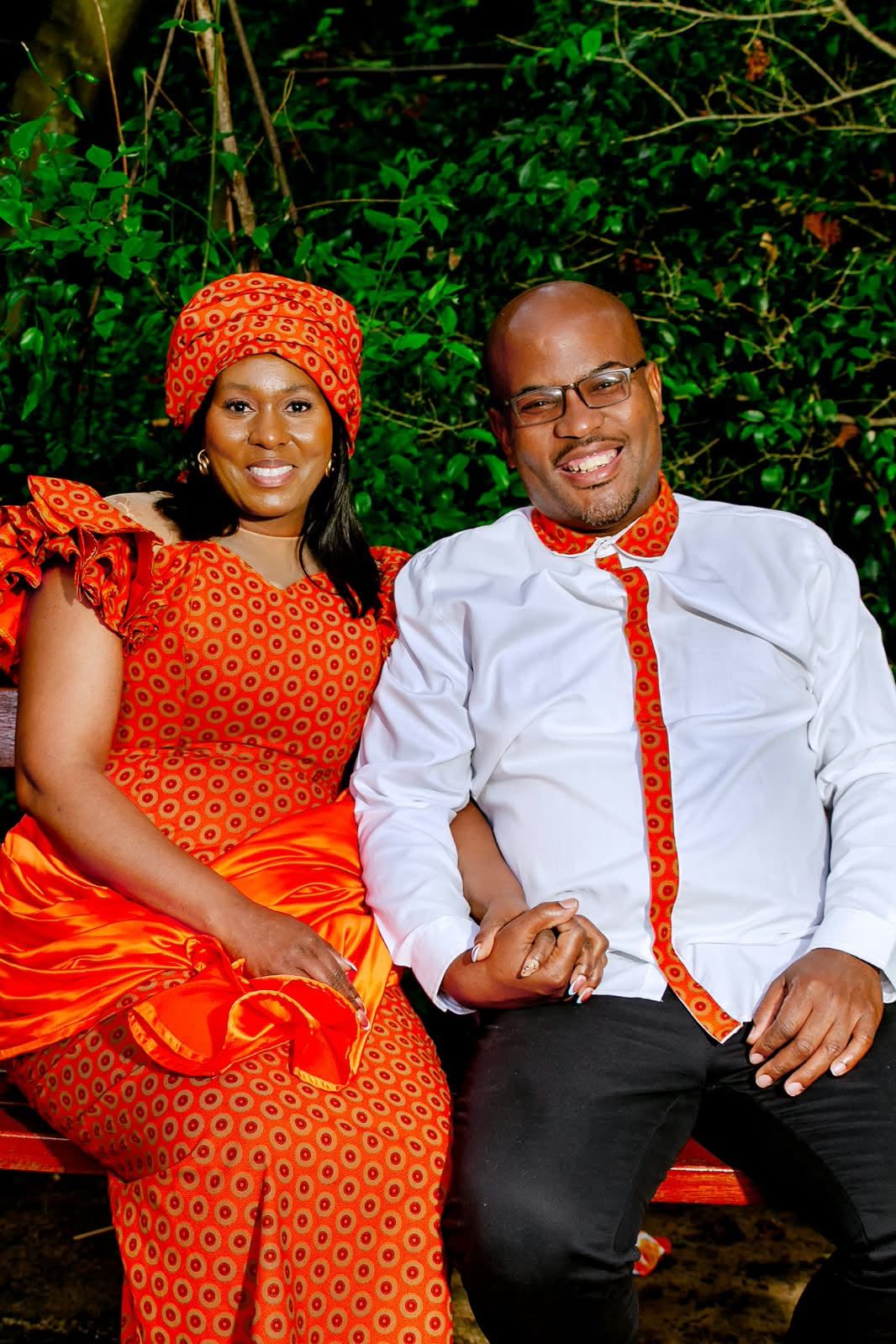 The couple seated on a red bench holding hands, she in an orange print dress and headwrap, he in a white shirt with matching orange trim